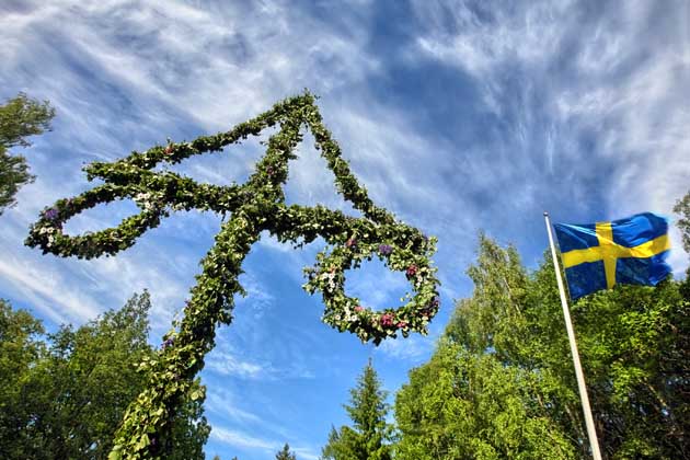 Mit Blumen geschmückter Maibaum und schwedische Flagge vor blauem Himmel an Mittsommer in Schweden