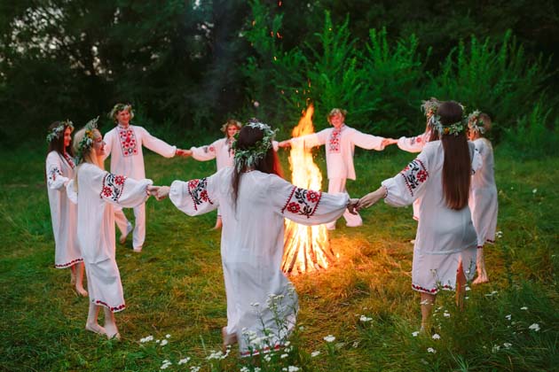 Junge Menschen mit Blumenkränzen im Haar tanzen an Mittsommer in weißen Kleidern, die an den Ärmeln bunt bestickt sind, im Kreis um ein Lagerfeuer im Wald herum