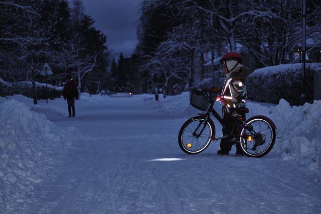 Ein Kind mit Fahrrad, Helm und vielen Reflektoren an seiner Kleidung steht in der Dämmerung im schneebedeckten Wald.