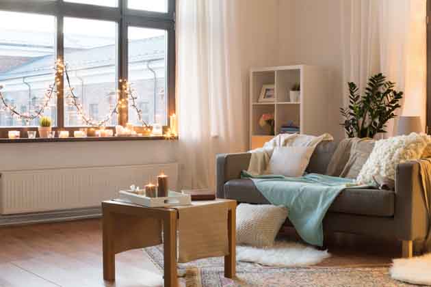 Ein gemütliches Wohnzimmer mit Sofa, Sofakissen, Regal, kleinem Tisch mit Kerzen und Lichterkette am Fenster.
