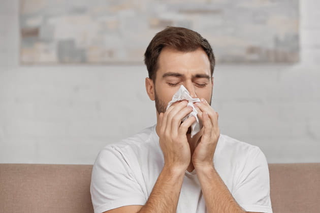 Blonder Mann mit Allergie schnaubt sich zu Hause auf dem Sofa sitzend die Nase mit einem Taschentuch