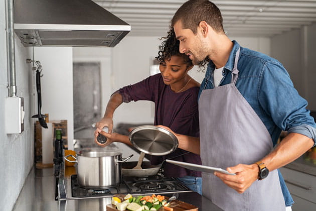Dampf vom Kochen wird durch die Dunstabzugshaube nach draußen geleitet, während eine junge Frau Salz in den Topf gibt und der Mann neben ihr ein Schneidebrett sowie ein Kochtopfdeckel in der Hand hält