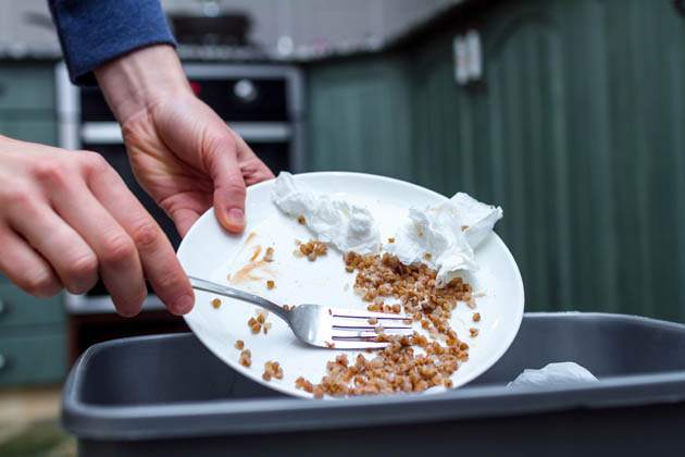 Nahaufnahme der Hände einer Person, die mit einer Gabel die Lebensmittelabfälle und Servietten von einem Teller in die Mülltonne schieben  