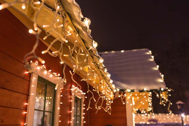 Das Hausdach eines roten Holzhauses ist mit Schnee bedeckt und hängenden Lichterketten geschmückt.