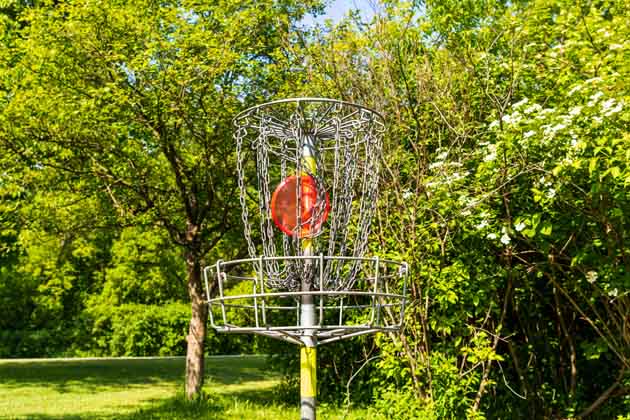 Eine orangenes Discgolf-Frisbee hängt im Netz des Fangkorbs - umgeben von grünen Bäumen und Gebüsch.