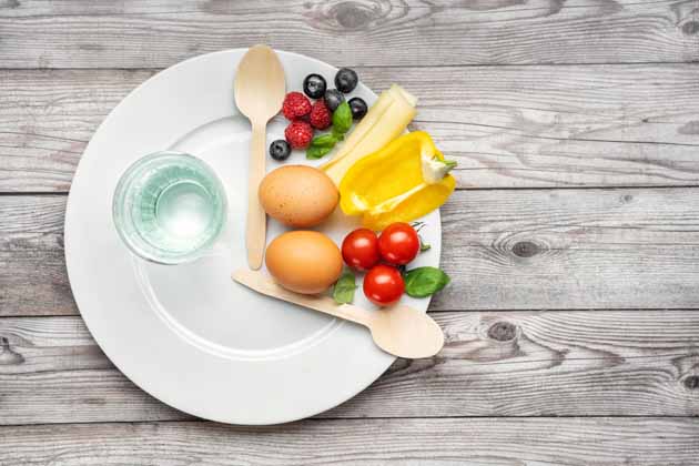 Ein Glas Wasser steht auf einem weißen Teller. Zwischen zwei Holzlöffeln auf dem Teller liegen Obst, Gemüse und Eier. Der Hintergrund ist ein grauer Holztisch.