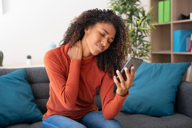 Eine Frau auf einer Couch mit Handy in der Hand hat Nackenschmerzen.