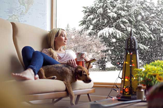 Eine Frau sitzt mit ihrem Hund entspannt auf der Couch und genießt den winterlichen Ausblick aus ihrem Fenster.