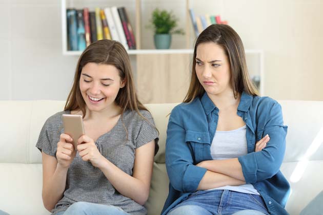 Zwei junge Frauen sitzen auf einer Couch. Eine guckt auf ihr Smartphone, was der anderen gar nicht gefällt.