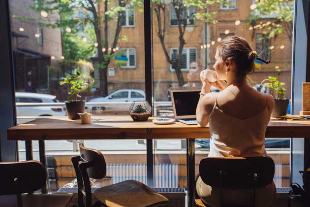 Eine junge Frau sitzt in einem Café am Fenster und arbeitet digital, während sie Urlaub macht (Workation).