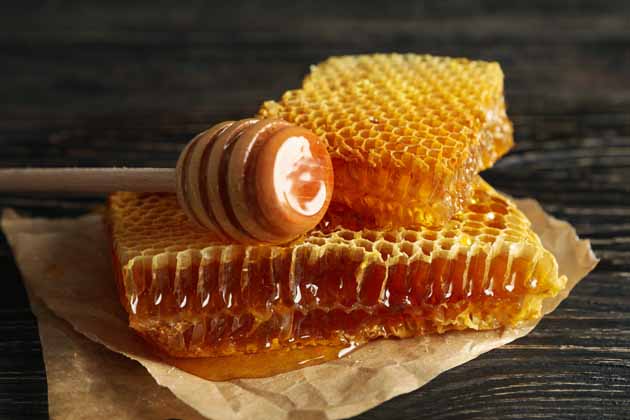 Zwei Stücke Honigwabe liegen mit einem Honiglöffel auf Backpapier auf einem dunklen Holztisch.