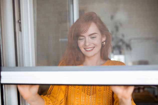 Eine Frau mit roten Haaren und orangenem Pullover schließt das Fenster mit Fliegengitter.