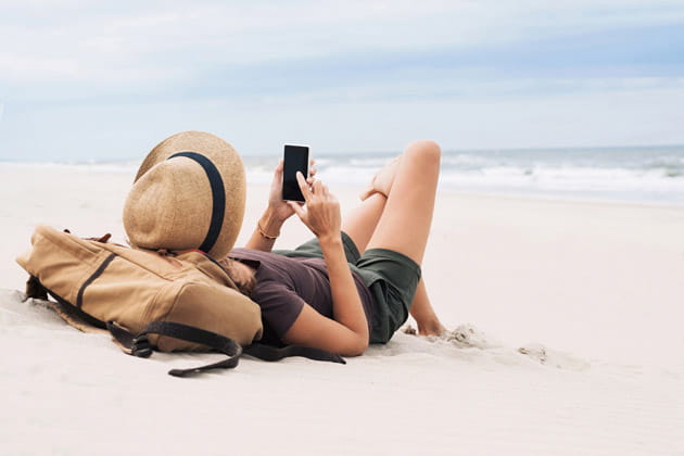Eine Person mit Sonnenhut und Rucksack liegt mit Smartphone am leeren Sandstrand.