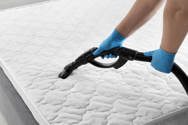 Eine Person mit blauen Gummihandschuhen saugt eine weiße Matratze ab.
