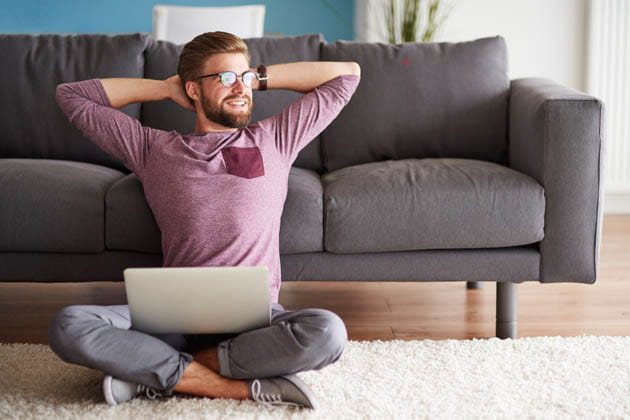 Ein junger Mann sitzt mit Laptop auf dem Teppich vor seinem Sofa und freut sich.
