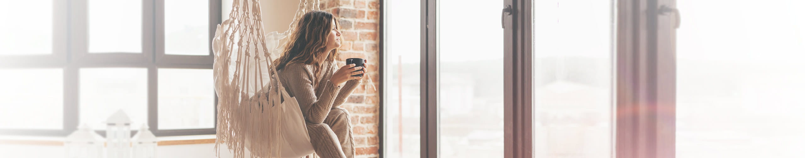 Frau steht mit Kaffeetasse nachdenklich vorm Fenster