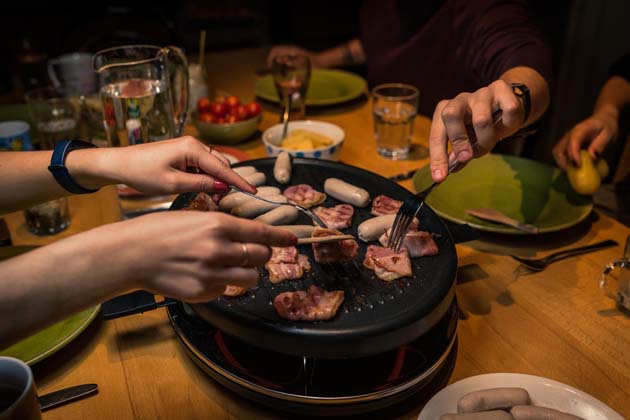 Mehrere Personen essen an einem Tisch Raclette und pieksen mit ihren Gabeln ins Fleisch auf dem Grill.